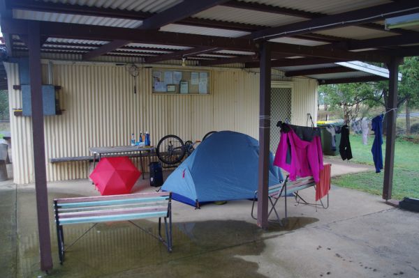 Small blue tent set up in outdoor covered cement floored space, open red umbrella, clothes on line