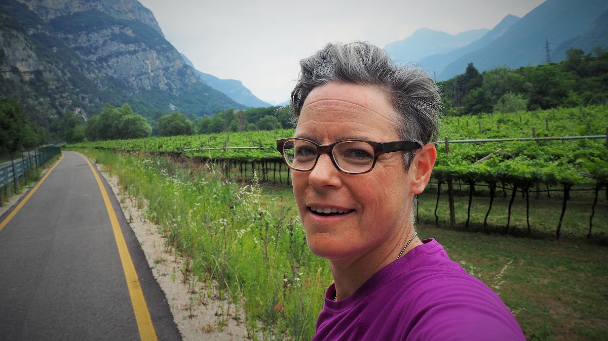 Selfie of woman bicycling in Italy - joyous woman. Cycleway, vineyards, and mountains in background